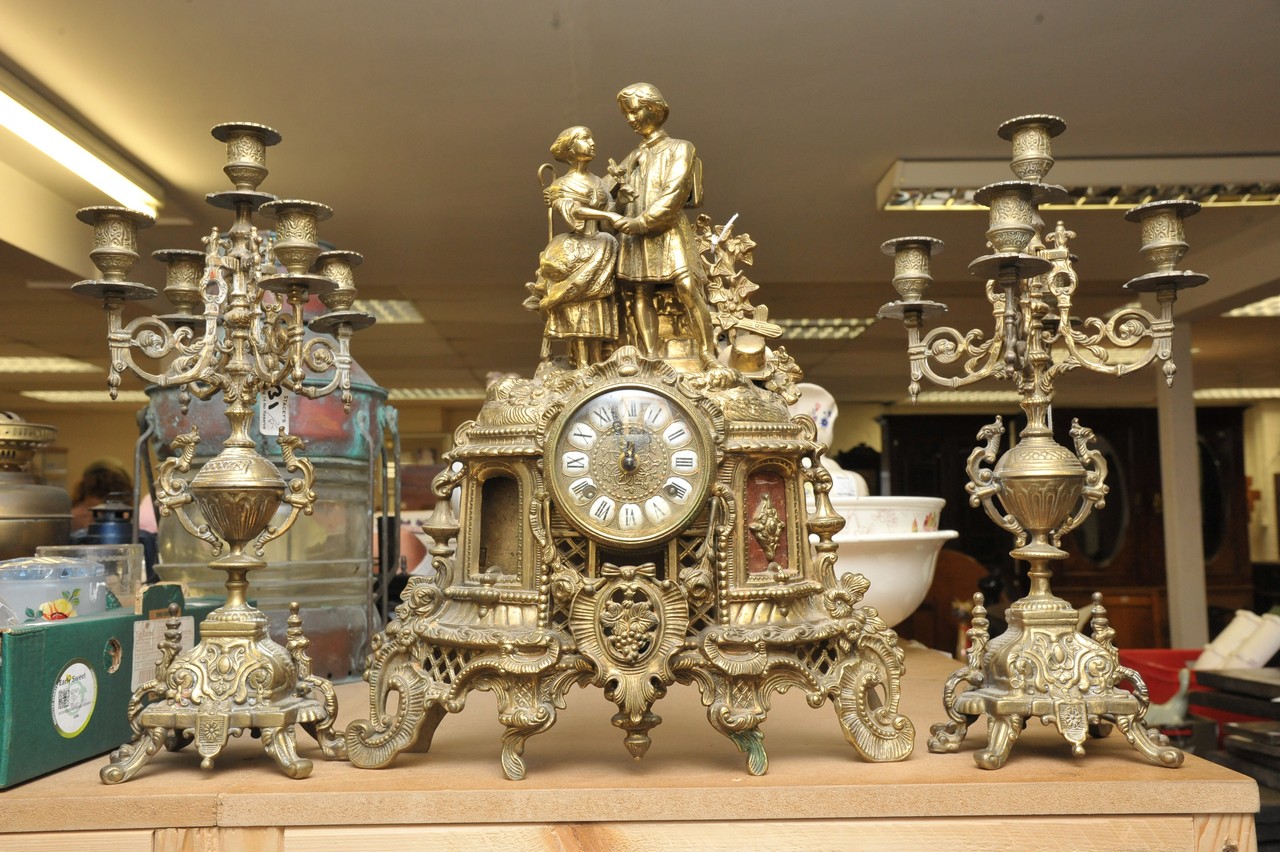 A cast metal clock garniture surmounted by figures with enamelled Roman Numeral dial