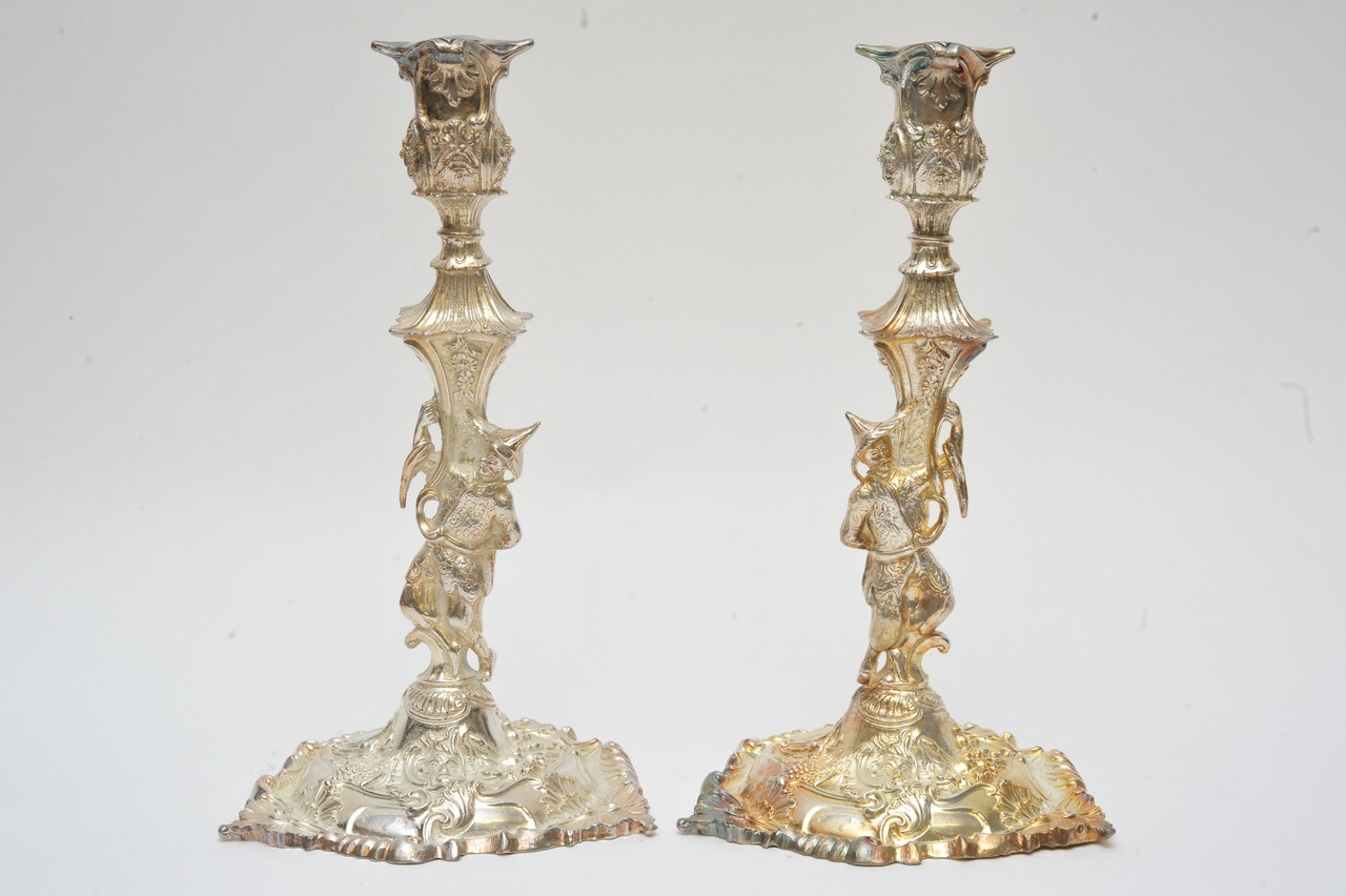 A pair of ornate silver plated candlesticks supported by figures with profuse raised scrolling