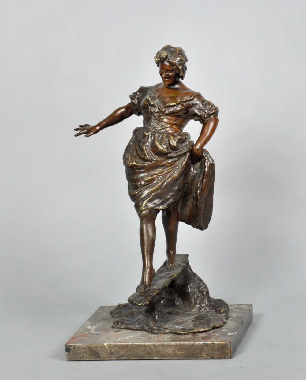 A late 19c cast bronze figure of a lady raising her skirt and negotiating a footbridge, signed