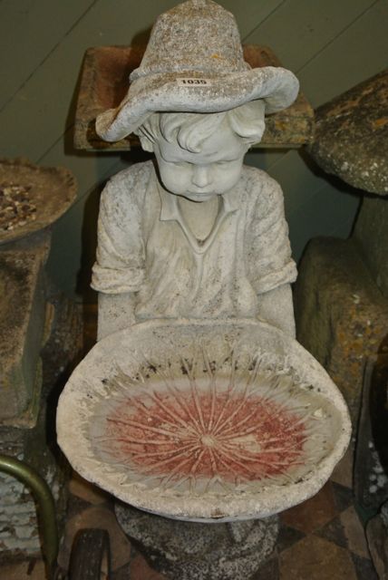 A weathered cast composition stone bird bath in the form of a boy figure in standing pose wearing a