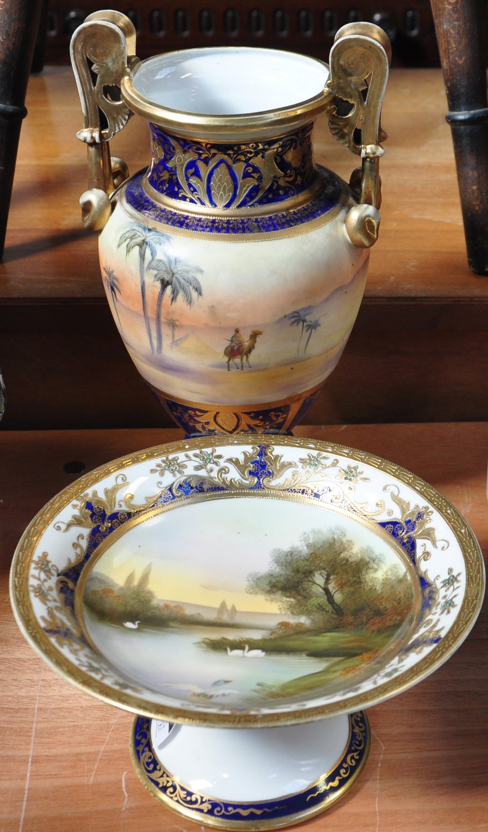 A Noritake hand painted gilt heightened comport decorated with swans on a tranquil river, diameter