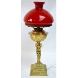 An ornate brass oil lamp with red glass shade, converted to electricity, height 67cm.