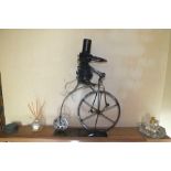 A novelty metal sculpture of a be-hatted bird riding a penny farthing, on plain rectangular plinth