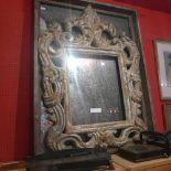 A large looking glass with heavy cast iron scolling leafy frame within square distressed pine frame