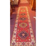 A Persian hand knotted Kazak runner the red field having central lozenge medallions in a multi