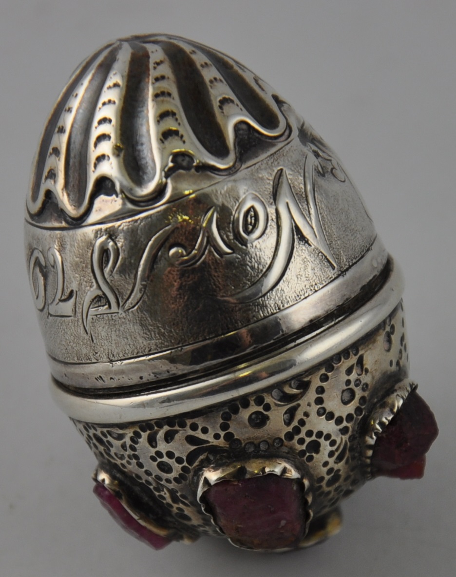 A white metal egg-shaped trinket box, set with uncut gemstones, possibly ruby, and inscribed