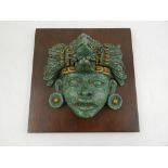 A South East Asian gilt and green wall ornament depicting the face of a deity, mounted on a wooden