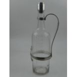 A clear glass and silver mounted whisky bottle, etched with measure divisions, within a white