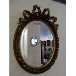 An oval bevelled wall mirror in a gilt frame with ribbon tie and leaf scroll decoration and a