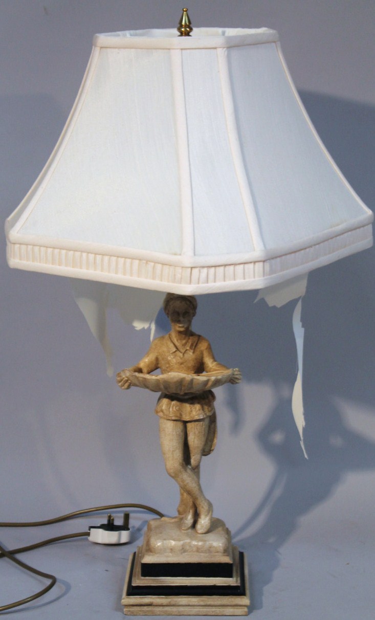 A resin table lamp, with white material shade on brass style column fronted by a figure holding a