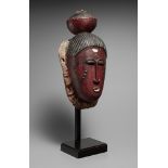 A BAULE MASK  The oval face painted red, black and white, the incised coiffure surmounted by a