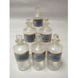 A set of six clear glass pharmaceutical bottles with stoppers, all with blue chemical labels with