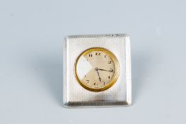 A near square engine turned silver easel clock case, the clock having a circular dial with brass