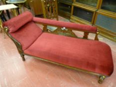 An Edwardian oak chaise longue with wine coloured upholstery, carved decoration raised on turned