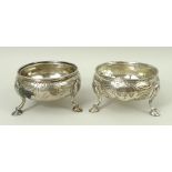 A pair of Victorian silver circular salts with foliate and rococo engraving raised on three hoof