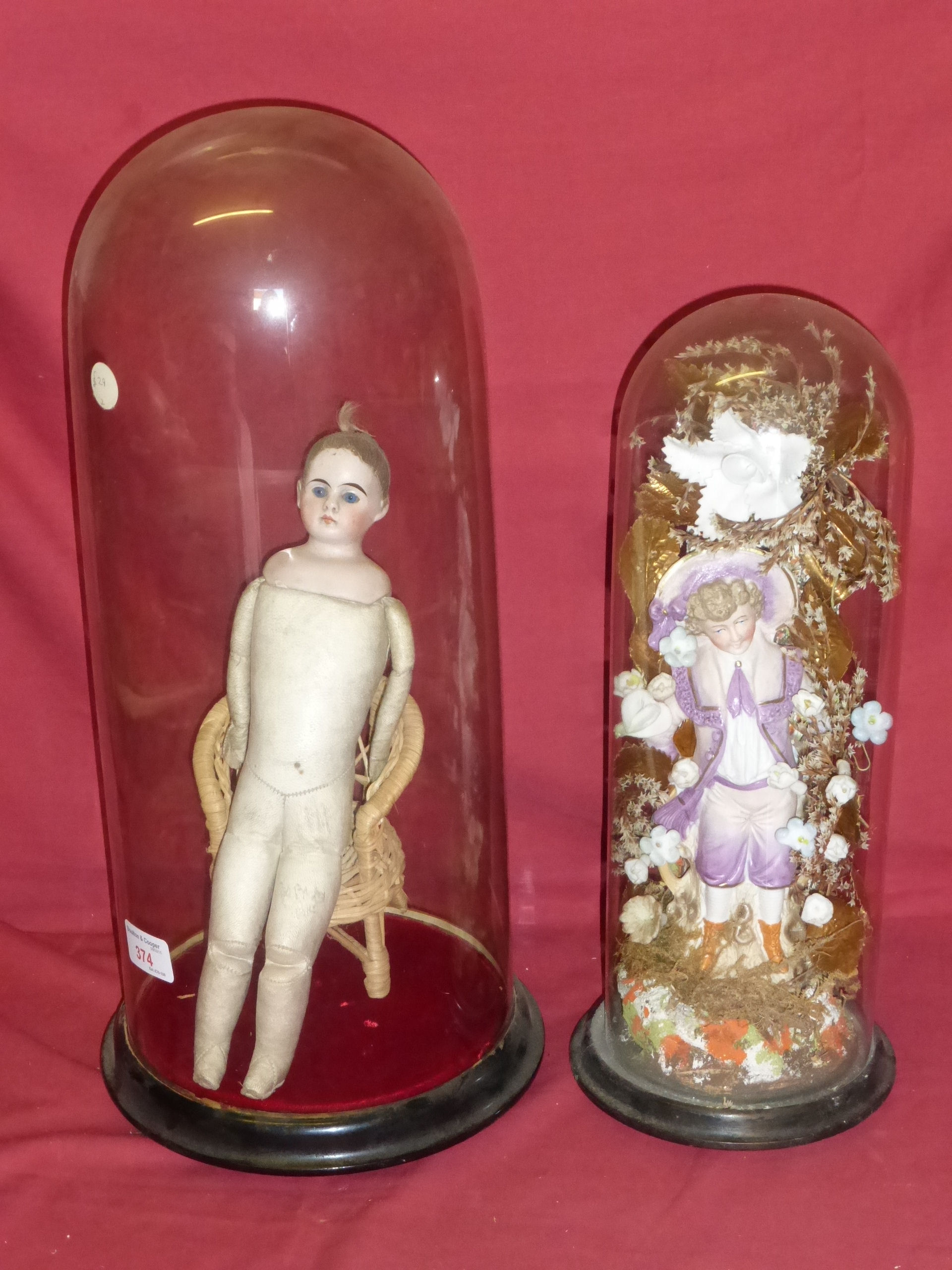 A small bisque head and leather covered Doll sitting in a wicker chair and under a glass dome shade,