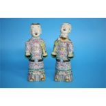 A pair of Chinese figures, each holding a vase on a mauve ground, red painted marks to the underside