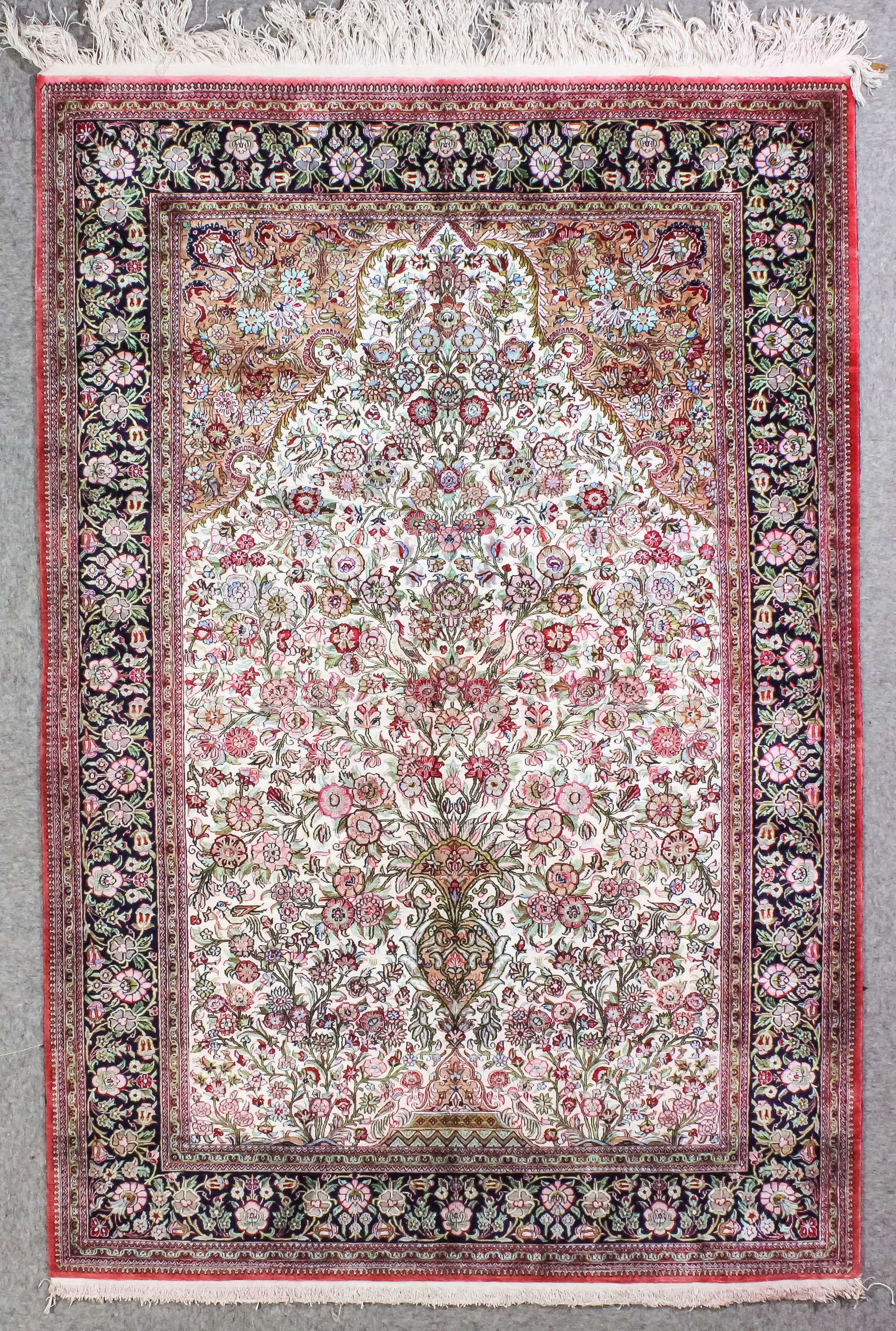 A Qum silk prayer rug woven in colours with a central mihrab filled with vase of flowers and birds
