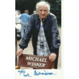 Michael Winner signed 8 x 6 colour photo standing behind his director’s chair. Good condition