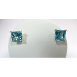 A pair of blue topaz single stone earstuds, each post headed by a princess cut vivid blue topaz in