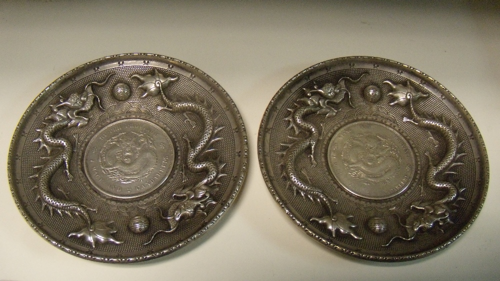 A pair of early 20th century silver coin inset dishes, the dishes cast and chased with two dragons