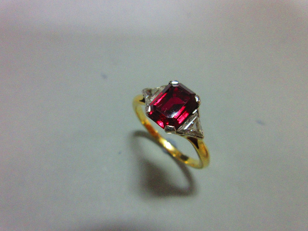 A ruby and diamond three stone ring, the emerald cut ruby four claw set between two trilliant cut