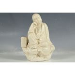 A Chinese cream statue, study of a young Buddhist scholar, seated upon a rock with books to his