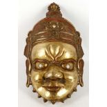 A Tibetan 19th Century brass god mask, red and gilt decoration to headdress, cloth internal padding,