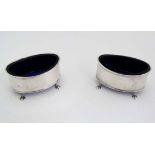 A pair of silver oval salt cellars with blue glass liners and standing on four outswept feet