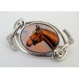 A white metal brooch with central ceramic cabochon with horse head decoration within a scrolling