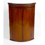 A bow-fronted wall-hanging mahogany corner cupboard. Having banded decoration to the double front