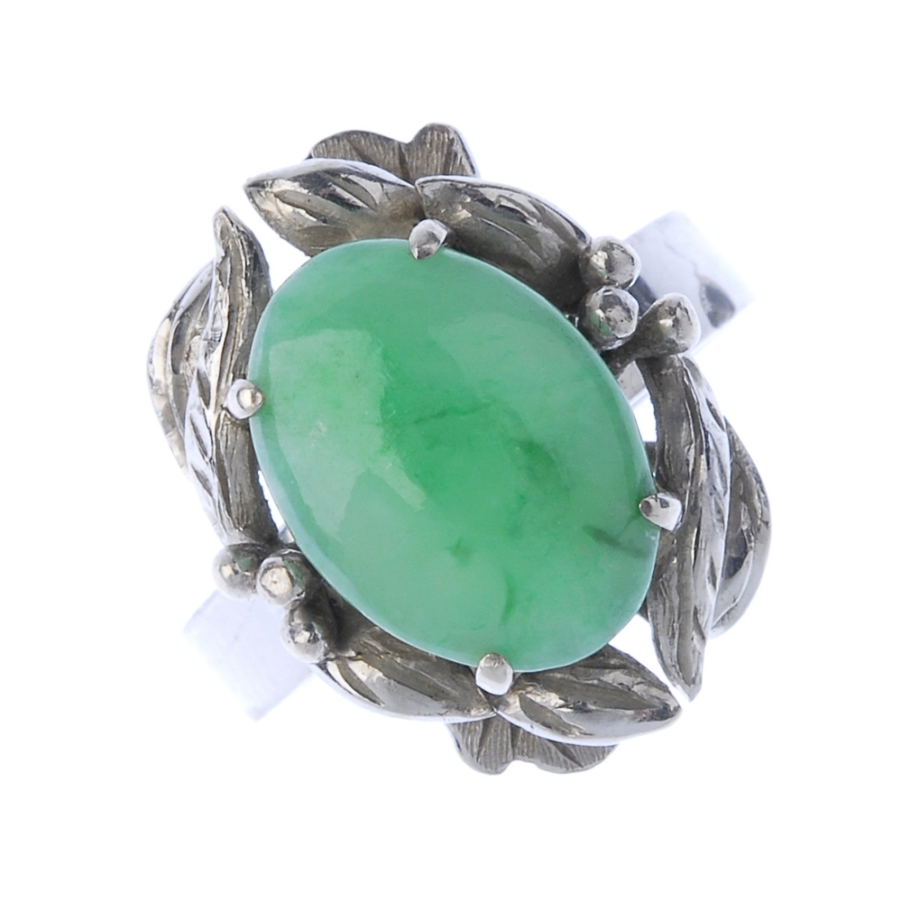 A jadeite dress ring. The oval jadeite cabochon, within a foliate surround, to the plain band.