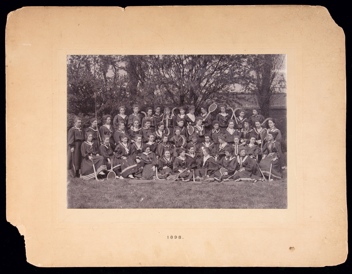 A photograph of a group of 45 schoolgirls holding tennis racquets and hockey sticks printed by W.