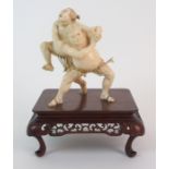 A Japanese ivory okimono of two sumo wrestlers in combat standing on a carved wood stand, the