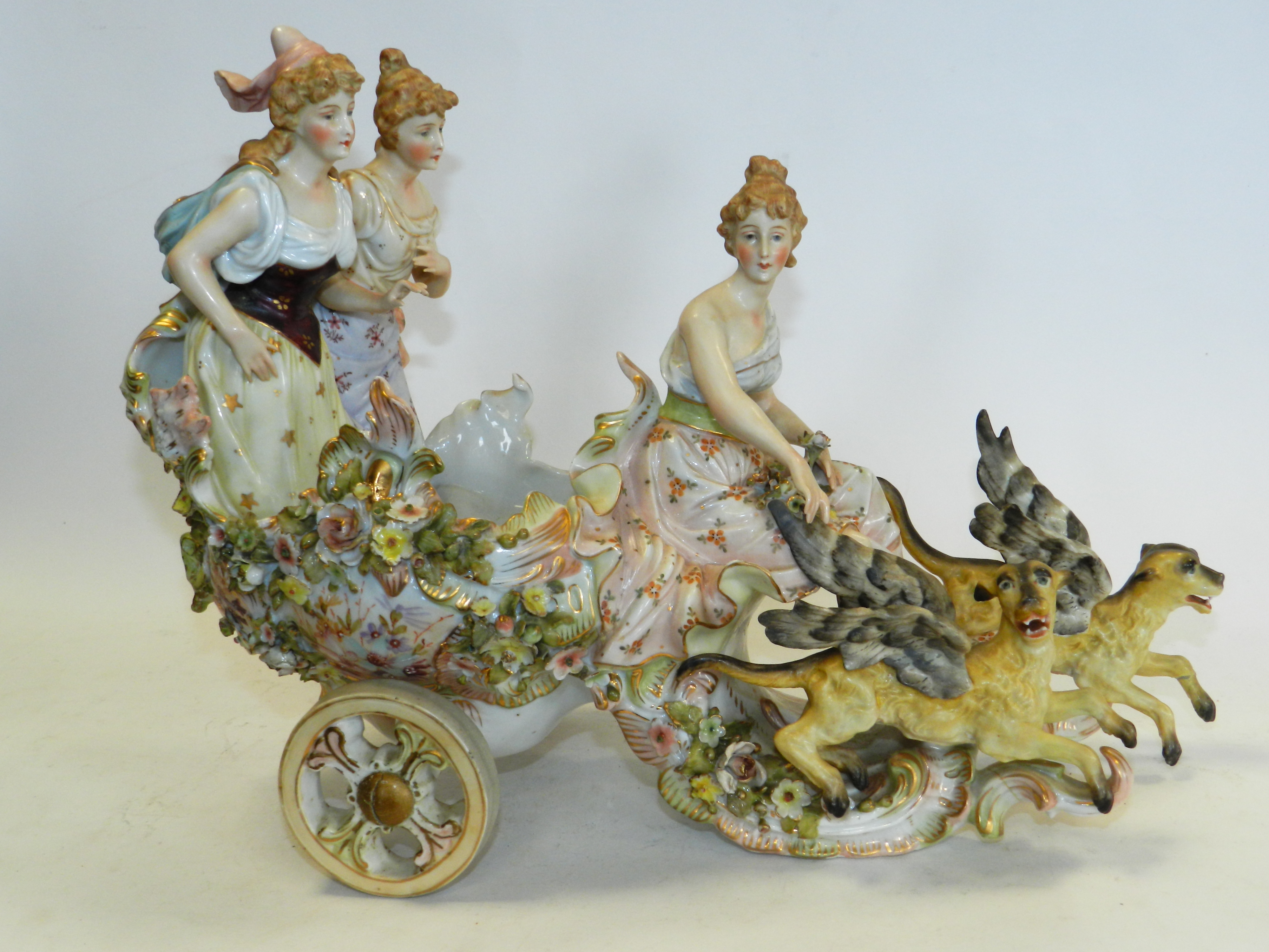 A Sitzendorf figural group of three ladies riding a chariot, pulled by two winged beasts (a.