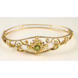 AN EDWARDIAN GOLD BANGLE, the open frame centred by a circular facet cut peridot within a border