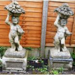 A PAIR OF RECONSTITUTED STONE FIGURES depicting cherubs holding a basket of fruit aloft, on square