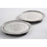 TWO VERY SIMILAR GEORGE III ENGRAVED OVAL TEA POT STANDS with bracket feet & a coat of arms in the