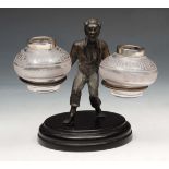 A BRONZE DESK STAND in the form of a figure holding two glass inkwells on a circular base, 10cm