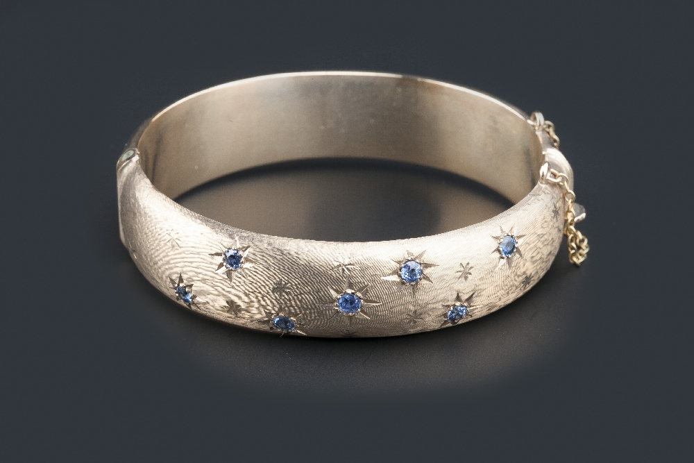 A 9CT GOLD AND SAPPHIRE SET BANGLE, of hinged oval design, star set with graduated circular-cut