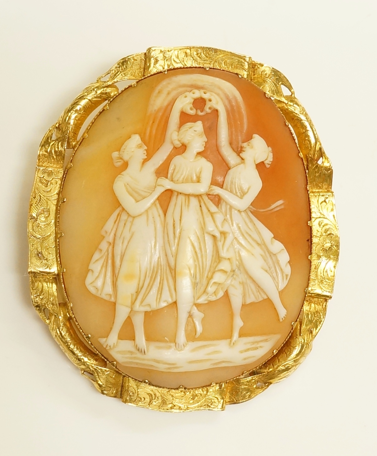 An oval shell cameo brooch the panel cut in relief with three graces within a 14ct gold foliate