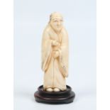 A Japanese carved ivory okimono raised on a carved hardwood plinth and formed as a man wearing