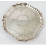A silver salver of shaped square form, with engraved decoration and raised on four scroll feet,