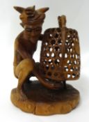 A Balinese carved wood figure of a seated man capturing a cockerel under a cage, 27cm