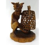 A Balinese carved wood figure of a seated man capturing a cockerel under a cage, 27cm
