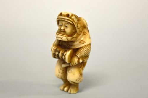 A JAPANESE IVORY NETSUKE, figurine dressed, in a Shi Shi dog mask banging a drum