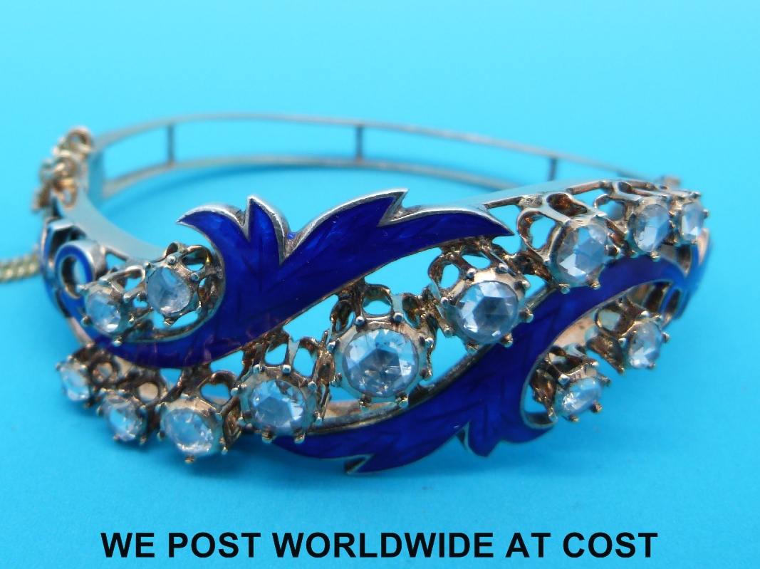 A yellow metal bangle set with 13 rose cut diamonds and blue enamel in a scrolling foliate design