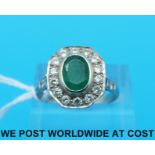 A white metal ring marked 18ct set with an oval cut emerald surrounded by round brilliant cut