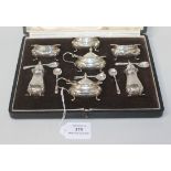 A George V silver seven piece condiment set of cushion form with scroll rims, comprising three