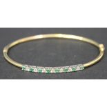 A 9ct gold, emerald and diamond set oval hinged bangle, the front mounted with a row of circular cut